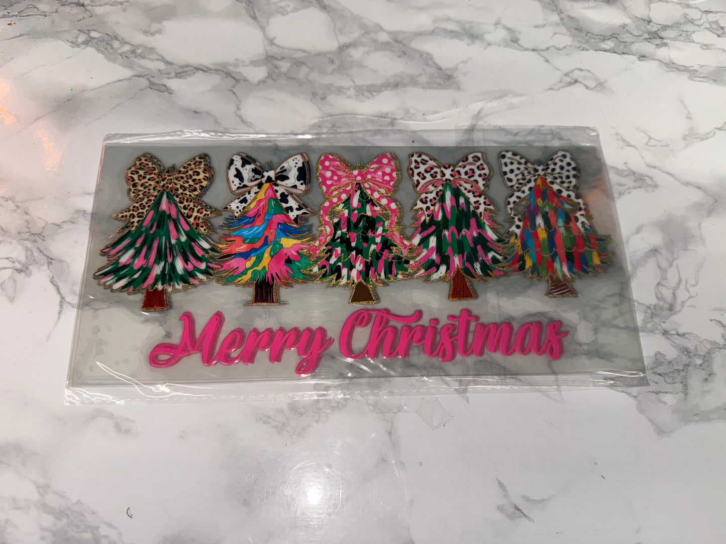 Decorative Christmas tree figurines with colorful patterns on a clear plastic card with 'Merry Christmas' text, placed on a marble surface.
